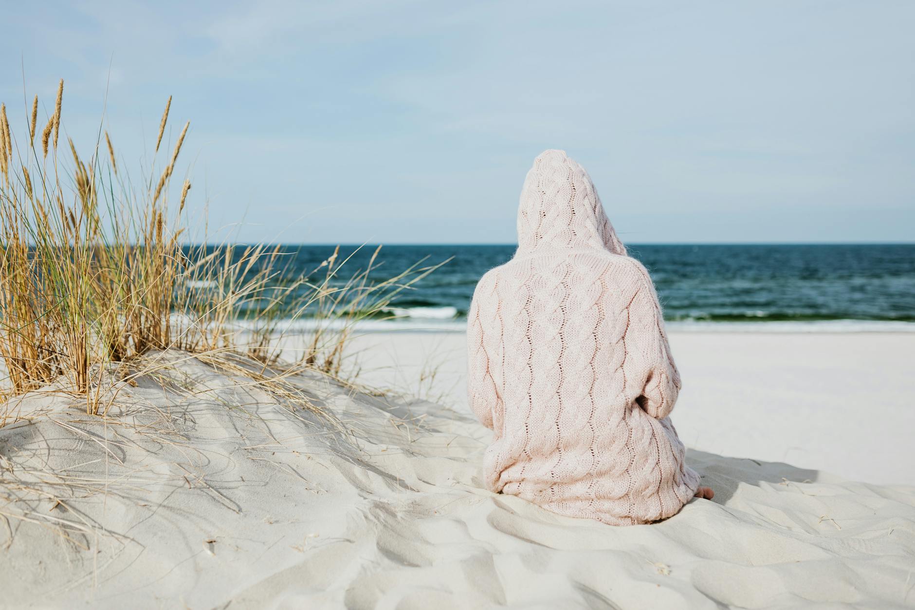 person wearing hoodie sitting on white sand near body of water