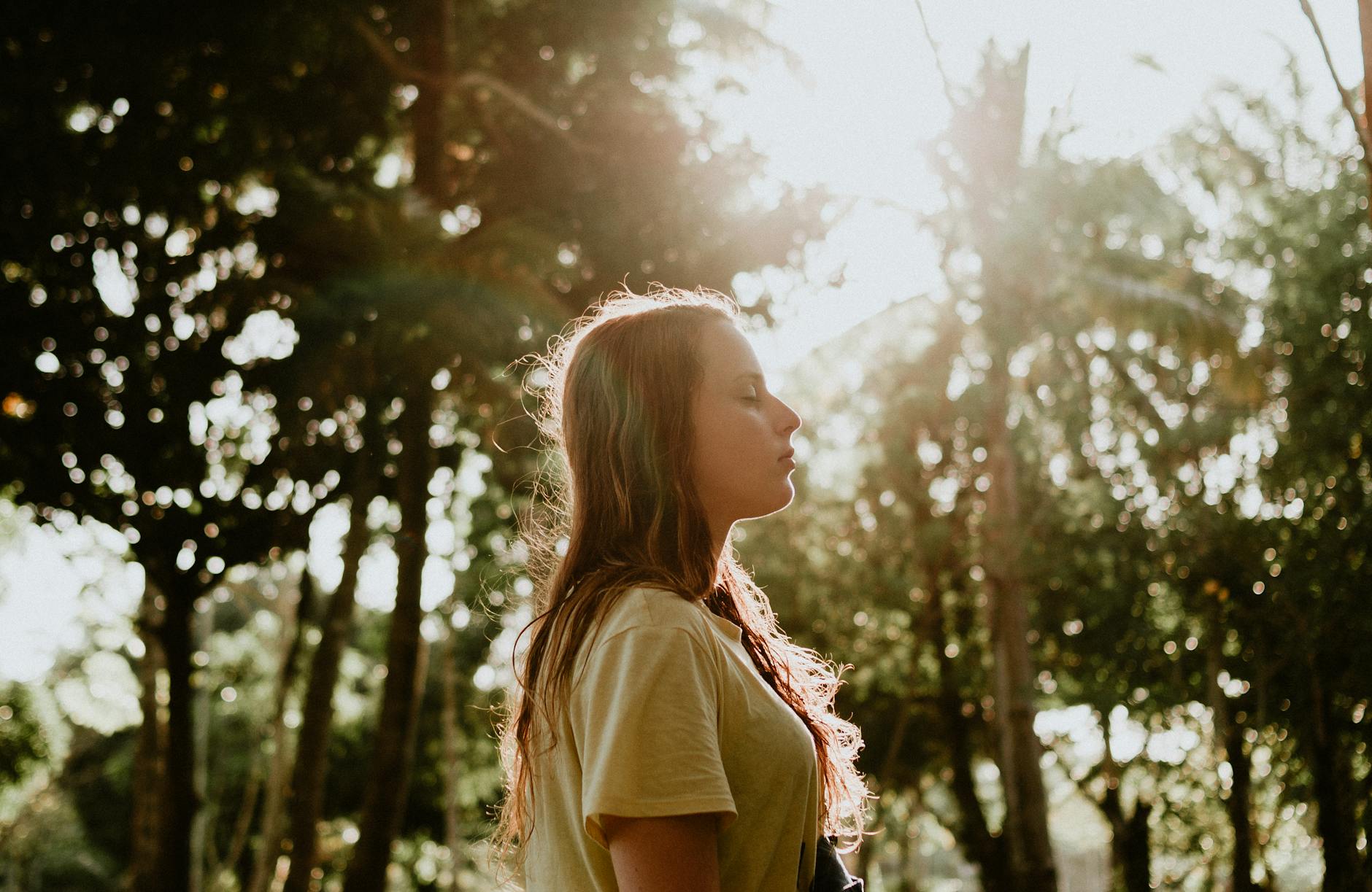 side view portrait photo of woman in yellow t shirt standing with her eyes closed with trees in the background