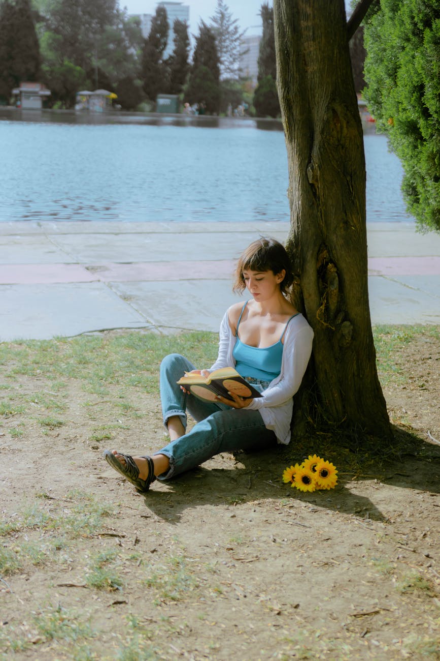 woman sitting leaning against a tree by the water and reading a book