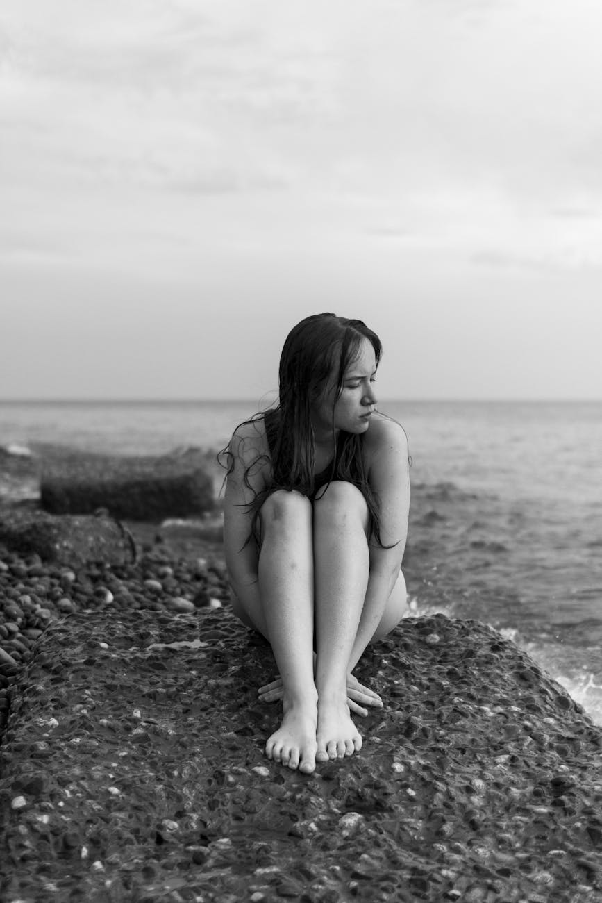 young naked woman sitting curled up on stony beach