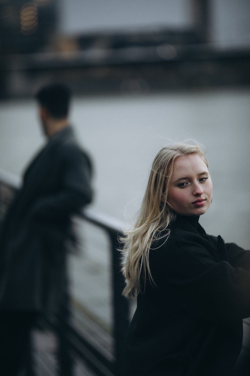 blonde woman in black coat standing by railing on street